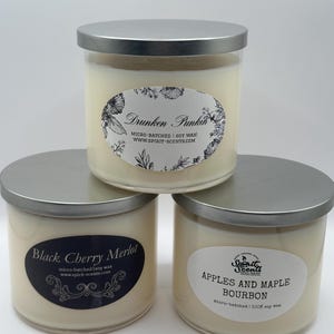 May include: Three glass candle jars with silver lids. The candles are white and have black labels with the names "Drunken Punkin", "Black Cherry Merlot", and "Apples and Maple Bourbon". Each label includes the text "micro-batched soy wax" and a website address.