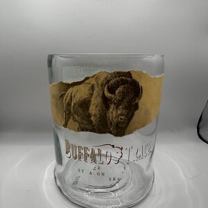 May include: A clear glass tumbler with a brown and tan label featuring a bison illustration and the words "Buffalo Trace". The glass has a wide base and a straight, cylindrical shape.