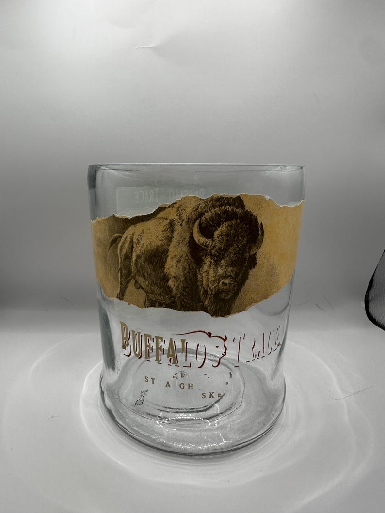 May include: A clear glass tumbler with a brown and tan label featuring a bison illustration and the words "Buffalo Trace". The glass has a wide base and a straight, cylindrical shape.