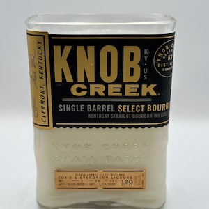 May include: A clear glass bottle with a white label that reads "Knob Creek Single Barrel Select Bourbon Kentucky Straight Bourbon Whiskey". The label is gold and black with the words "Knob Creek" in gold letters. The bottle is sitting on a white surface.