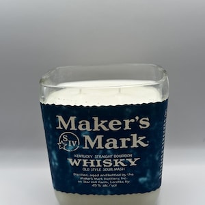 May include: A square glass candle with a white wax interior. The candle has a blue label with the text "Maker's Mark Whisky" and other details about the product. The candle is a bourbon-themed item.