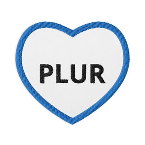 Può includere: Una toppa a forma di cuore blu con la scritta "PLUR" in lettere nere.
