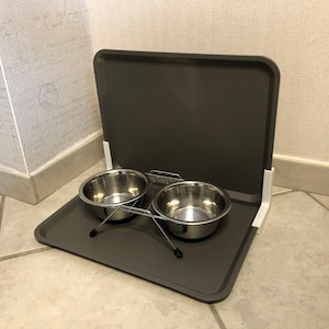 Könnte beinhalten: Ein grauer, faltbarer Futter- und Wassernapfständer für Haustiere mit zwei Schüsseln aus Edelstahl. Der Ständer hat weiße Kunststoffstützen und einen Metallrahmen.