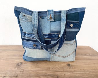 May include: A patchwork denim tote bag in various shades of blue. The bag features multiple denim patches, pockets, and a button closure. The handles are made of the same denim material. The bag is sitting on a wooden surface.