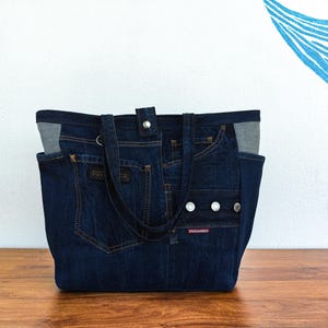Könnte beinhalten: Eine dunkelblaue Tragetasche aus recyceltem Denim mit mehreren Taschen, silberfarbenen Metallknöpfen, einem zentralen Druckknopfverschluss und einem roten rechteckigen Etikett mit dem weißen Schriftzug DSQUARED2 auf der Vorderseite.