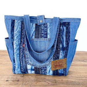 Może przedstawiać: Niebieska torba typu tote z dżinsu z patchworkowym wzorem. Torba posiada dwie zewnętrzne kieszenie, zapięcie na guzik i dżinsowe uchwyty. Brązowa skórzana metka jest przyszyta z przodu. Torba stoi na drewnianej powierzchni.