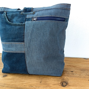 K&ouml;nnte beinhalten: Eine blaue Denim-Tragetasche aus recycelten Jeans. Die Tasche hat einen Rei&szlig;verschluss, eine Vordertasche und besteht aus verschiedenen Blaut&ouml;nen. Die Tasche ist rechteckig und wirkt robust.