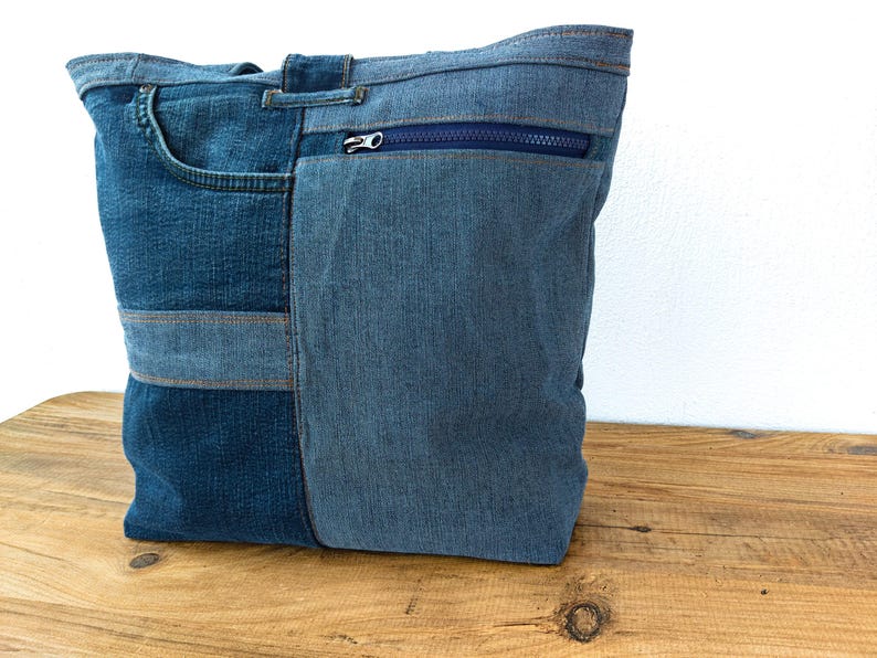 K&ouml;nnte beinhalten: Eine blaue Denim-Tragetasche aus recycelten Jeans. Die Tasche hat einen Rei&szlig;verschluss, eine Vordertasche und besteht aus verschiedenen Blaut&ouml;nen. Die Tasche ist rechteckig und wirkt robust.