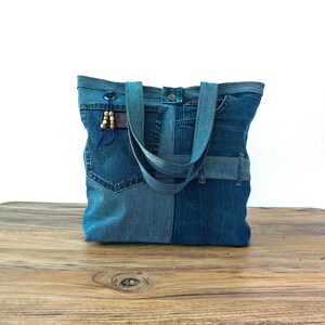 K&ouml;nnte beinhalten: Eine Denim-Tragetasche aus recycelten Jeans. Die Tasche hat ein Patchwork-Design mit verschiedenen Blaut&ouml;nen. Sie hat zwei Tr&auml;ger und eine kleine blaue Kordel mit Holzperlen. Die Tasche steht auf einer Holzoberfl&auml;che.