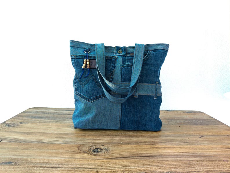 K&ouml;nnte beinhalten: Eine Denim-Tragetasche aus recycelten Jeans. Die Tasche hat ein Patchwork-Design mit verschiedenen Blaut&ouml;nen. Sie hat zwei Tr&auml;ger und eine kleine blaue Kordel mit Holzperlen. Die Tasche steht auf einer Holzoberfl&auml;che.