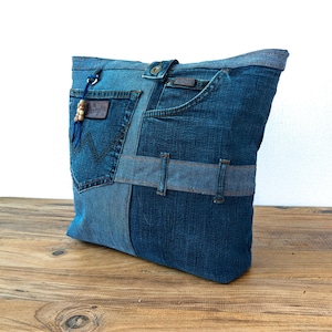 K&ouml;nnte beinhalten: Eine Denim-Tragetasche aus recycelten Jeans. Die Tasche zeigt verschiedene Blaut&ouml;ne von Denim, mit Taschen und G&uuml;rtelschlaufen, die in das Design integriert sind. Ein Lederaufn&auml;her und Holzperlen sorgen f&uuml;r Details.
