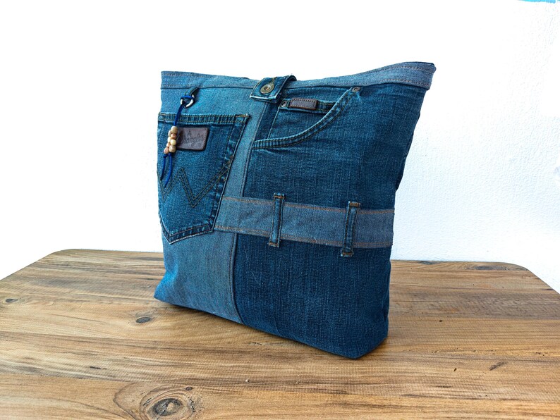 K&ouml;nnte beinhalten: Eine Denim-Tragetasche aus recycelten Jeans. Die Tasche zeigt verschiedene Blaut&ouml;ne von Denim, mit Taschen und G&uuml;rtelschlaufen, die in das Design integriert sind. Ein Lederaufn&auml;her und Holzperlen sorgen f&uuml;r Details.