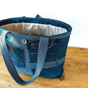 K&ouml;nnte beinhalten: Eine Denim-Tragetasche aus recycelten Jeans. Die Tasche hat mehrere Taschen, einen Knopfverschluss und stabile Tr&auml;ger. Der Innenraum ist mit einem hellen Stoff gef&uuml;ttert. Die Tasche ist f&uuml;r den Transport von Alltagsgegenst&auml;nden konzipiert.
