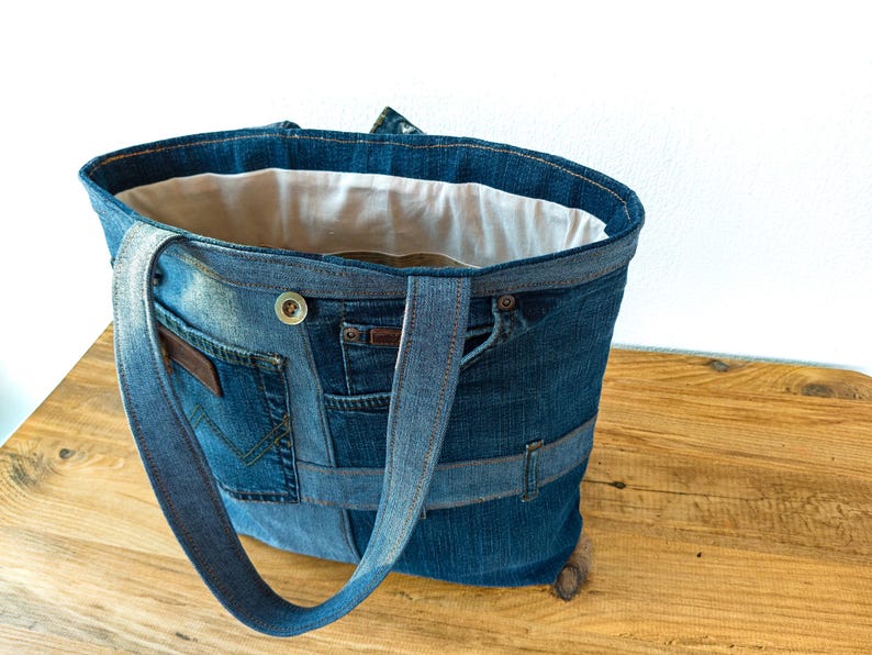 K&ouml;nnte beinhalten: Eine Denim-Tragetasche aus recycelten Jeans. Die Tasche hat mehrere Taschen, einen Knopfverschluss und stabile Tr&auml;ger. Der Innenraum ist mit einem hellen Stoff gef&uuml;ttert. Die Tasche ist f&uuml;r den Transport von Alltagsgegenst&auml;nden konzipiert.