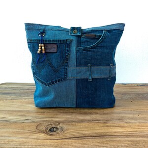 K&ouml;nnte beinhalten: Eine Denim-Tragetasche aus recycelten Jeans. Die Tasche hat ein Patchwork-Design mit verschiedenen Blaut&ouml;nen, einschlie&szlig;lich Taschen und G&uuml;rtelschlaufen. Ein kleines Lederpatch und Holzperlen f&uuml;gen Details hinzu. Die Tasche wird auf einer Holzoberfl&auml;che pr&auml;sentiert.