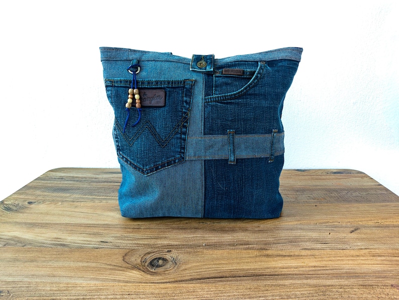 K&ouml;nnte beinhalten: Eine Denim-Tragetasche aus recycelten Jeans. Die Tasche hat ein Patchwork-Design mit verschiedenen Blaut&ouml;nen, einschlie&szlig;lich Taschen und G&uuml;rtelschlaufen. Ein kleines Lederpatch und Holzperlen f&uuml;gen Details hinzu. Die Tasche wird auf einer Holzoberfl&auml;che pr&auml;sentiert.