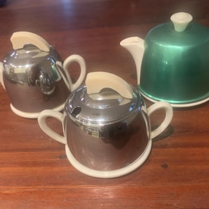 May include: Three small teapots and a sugar bowl on a wooden surface. The teapots are silver and green, with white spouts, handles, and bases. The sugar bowl is silver with two handles and a lid. The lids and handles are a cream colour.