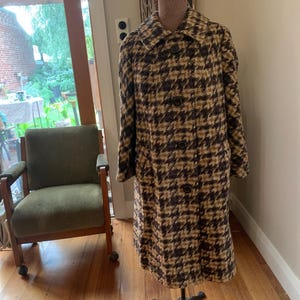 May include: A long, houndstooth patterned coat in shades of brown and black. The coat has a classic collar and three large buttons down the front. The coat is displayed on a mannequin in a room with a green chair and a window.