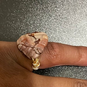 Könnte beinhalten: Ein goldener Ring mit einem herzförmigen Stein. Der Stein ist eine Mischung aus braunen, weißen und rosafarbenen Farben mit einem marmorierten Muster.
