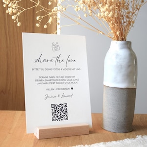 Könnte beinhalten: Eine weiße Karte mit dem Text "share the love" und einem QR-Code, gehalten von einem Holzständer. Rechts steht eine Keramikvase mit Trockenblumen. Der Text lädt Gäste ein, Fotos und Videos zu teilen.