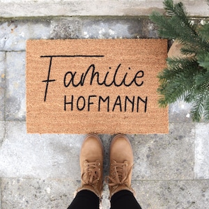 Eine braune Fußmatte aus Kokosfaser liegt auf grauem Stein. Auf der Fußmatte befinden sich die Worte Familie Hofmann in schwarzer Schrift.