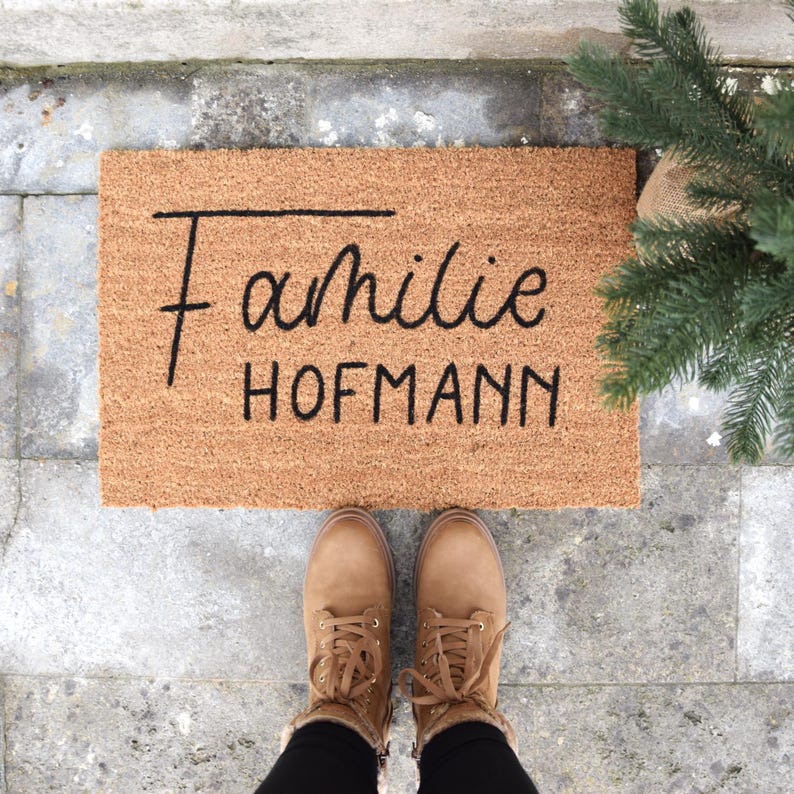 Eine braune Fußmatte aus Kokosfaser liegt auf grauem Stein. Auf der Fußmatte befinden sich die Worte Familie Hofmann in schwarzer Schrift.