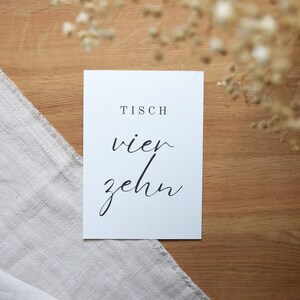 May include: White card with black text that reads "Tisch vier zehn". The card is on a wooden surface.