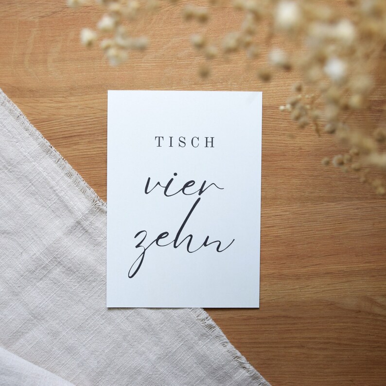 May include: White card with black text that reads "Tisch vier zehn". The card is on a wooden surface.