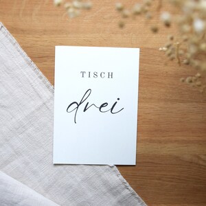 May include: White card with black text that reads "Tisch drei" on a wooden surface.