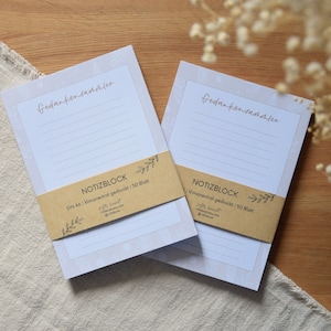 Könnte beinhalten: Zwei weiße Notizblöcke mit einem floralen Design und dem Text "Gedankensammler" auf der Vorderseite. Die Notizblöcke sind in braunes Papier eingewickelt, auf dem "NOTIZBLOCK" und "DIN A6 klimaneutral gedruckt | 50 Blatt" steht.