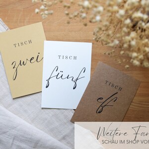 May include: Three table number cards with the text "Tisch" (German for table) and the numbers "zwei" (two), "f&uuml;nf" (five), and "elf" (eleven) in black script font. The cards are on a light brown wooden surface with a white linen cloth.