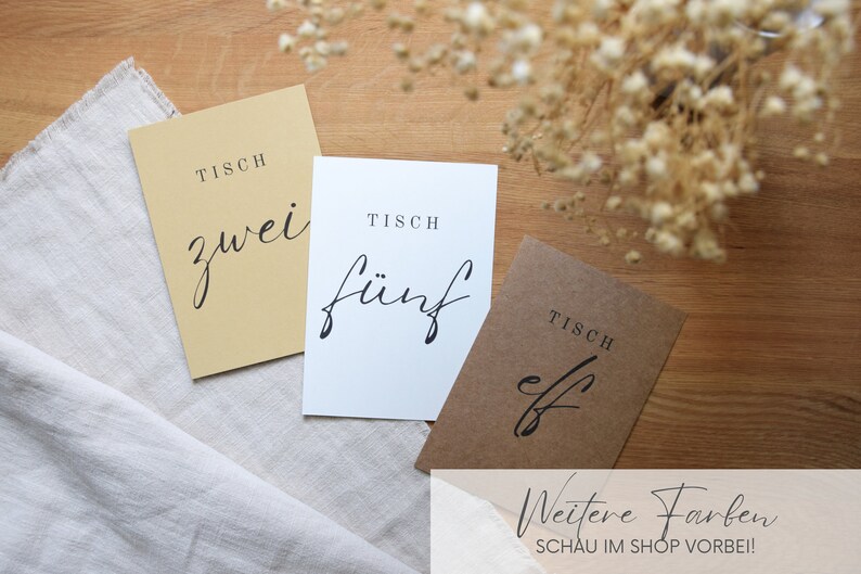 May include: Three table number cards with the text "Tisch" (German for table) and the numbers "zwei" (two), "f&uuml;nf" (five), and "elf" (eleven) in black script font. The cards are on a light brown wooden surface with a white linen cloth.