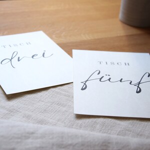 May include: Two white cards with black text. The first card says "Tisch drei" and the second card says "Tisch f&uuml;nf".