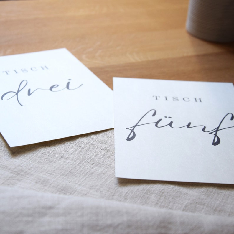 May include: Two white cards with black text. The first card says "Tisch drei" and the second card says "Tisch f&uuml;nf".