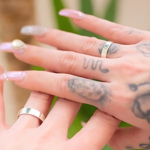 May include: Two smooth, polished silver bands worn on fingers. Each ring features a simple, flat, and unadorned design with a reflective metallic finish. The bands are uniform in width and color, showcasing a clean, minimalist aesthetic on the fingers.