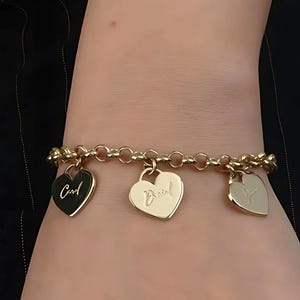 Puede incluir: Pulsera de cadena dorada con tres dijes en forma de corazón. Los dijes están grabados con las palabras "Cool", "Devil" y un símbolo de corazón.