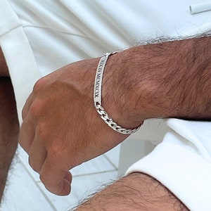 Könnte beinhalten: Ein silbernes Panzerarmband mit einem rechteckigen Schild, das mit römischen Ziffern graviert ist. Das Armband wird am Handgelenk getragen, wobei die Kettenglieder deutlich sichtbar sind. Der Hintergrund ist eine Person, die weiße Shorts trägt.