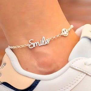 Puede incluir: Una tobillera de tono plateado que presenta una cadena delicada con un dije en letra cursiva que dice la palabra Smile y un pequeño dije circular de cara sonriente. La tobillera se coloca alrededor de un tobillo sobre una zapatilla blanca.
