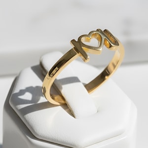 Op de afbeelding: Een goudkleurige ring met een uitgesneden ontwerp op de band dat de letters I, een hartvorm en de letter U vormt. De ring heeft een gepolijst, reflecterend oppervlak en een dunne, ronde band die aansluit op het centrale letterontwerp.
