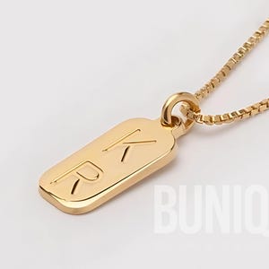 May include: A gold-toned necklace featuring a rectangular pendant with rounded corners. The pendant is engraved with the letters K and R. The pendant hangs from a delicate box chain. The text BUNIQE JEWELRY is visible in the lower right corner.