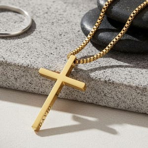May include: A gold-colored cross pendant necklace with the name "DAVID" engraved on the side. A silver ring is in the background. The necklace is on a gold chain. The cross is resting on a gray surface.
