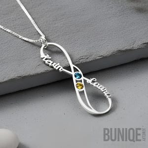 Puede incluir: Un collar con colgante de infinito en plata que presenta los nombres Kevin y Laura en letra cursiva. El colgante incluye dos pequeñas piedras preciosas circulares, una azul y otra amarilla, engastadas en el diseño de infinito en una cadena fina.