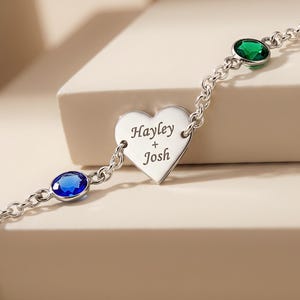 May include: A silver-toned chain bracelet featuring a central heart-shaped charm engraved with the text Hayley + Josh. The bracelet is accented with one round blue gemstone and one round green gemstone set into the chain links on either side.