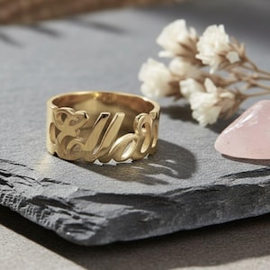 May include: A gold ring engraved with the name "Ella" in a cursive style. The ring is displayed on a dark grey slate, with white flowers and a pink crystal in the background. The ring is wide.