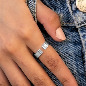 May include: A silver-toned open band ring featuring two flat, rectangular ends. Each end is engraved with a single capital letter, specifically the letters S and E, positioned on the finger against a background of blue denim fabric.