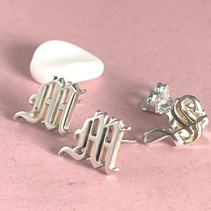 May include: A pair of silver earrings with a gothic letter 'M' design. The earrings are on a pink surface.