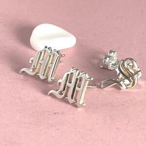 Könnte beinhalten: Ein Paar silberne Ohrstecker in Form des Buchstabens M in einer gotischen Schriftart, begleitet von einem separaten silbernen Ohrsteckerverschluss. Die metallisch silberne Oberfläche reflektiert das Licht und verleiht den Ohrsteckern Glanz.