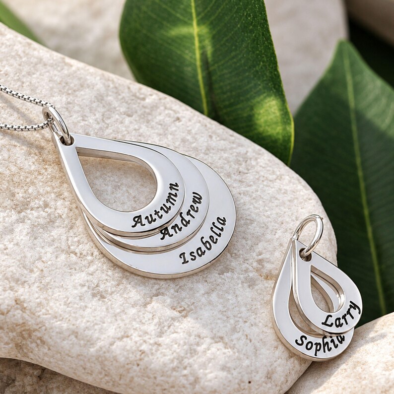 May include: A silver-toned necklace featuring three stacked teardrop-shaped pendants engraved with the names Autumn, Andrew, and Isabella, alongside a separate smaller charm with two teardrop pendants engraved with the names Larry and Sophia.