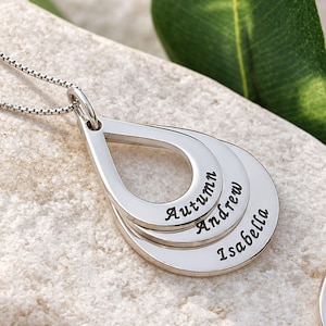 May include: A silver-toned necklace featuring three stacked, teardrop-shaped pendants. Each pendant is engraved with a name in a cursive script: Autumn, Andrew, and Isabella. The pendants are suspended from a delicate silver box chain.