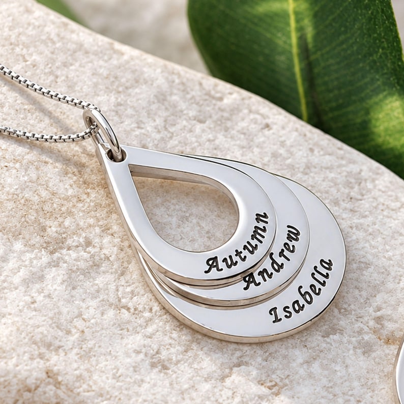 May include: A silver-toned necklace featuring three stacked, teardrop-shaped pendants. Each pendant is engraved with a name in a cursive script: Autumn, Andrew, and Isabella. The pendants are suspended from a delicate silver box chain.